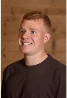 Porträt eines jungen Mannes mit kurzem, hellbraunem Haar, der lächelnd leicht nach rechts blickt. Er trägt ein dunkles T-Shirt, im Hintergrund ist eine warme Holzverkleidung zu sehen.