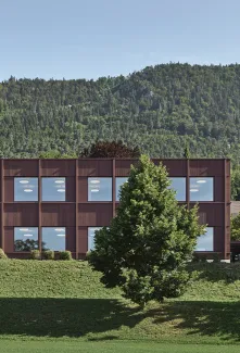 Aussenansicht eines modernen, zweigeschossigen Gebäudes mit dunkelbrauner Fassade und grossen, regelmässig angeordneten Fenstern. Das Gebäude steht in einer grünen Umgebung mit Wiese und Bäumen. Im Hintergrund erhebt sich ein bewaldeter Hügel unter klarem Himmel, was die ruhige, naturnahe Lage des Baus betont.