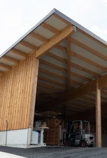 Aussenansicht einer grossen, offenen Lager- oder Maschinenhalle in Holzbauweise mit geneigtem Dach. Die Konstruktion aus sichtbaren Holzbalken ruht auf einem Betonsockel. Im Inneren der Halle sind ein Gabelstapler und gelagerte Materialien zu erkennen. Die Umgebung wirkt ländlich und funktional gestaltet.
