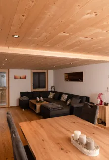 Innenansicht eines gemütlichen Wohnzimmers mit hellem Holzboden und sichtbarer Holzdecke. An der Wand hängt ein Fernseher über einem niedrigen Sideboard. Mehrere Holzfenster geben den Blick in eine bewaldete Landschaft frei. Rechts führt eine Glastür in einen angrenzenden Raum, der Wohnbereich ist warm und wohnlich gestaltet.