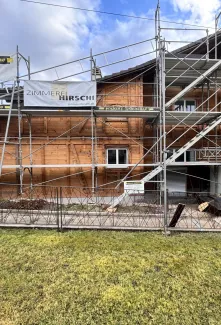 Holzhaus im Bau mit eingerüsteter Fassade und Baustellenbanner der Zimmerei Hirschi unter blauem Himmel.
