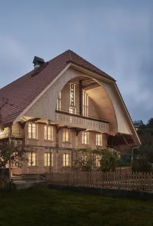 Traditionelles Holzhaus im Emmental in der Abenddämmerung, mit geschwungenem Giebeldach und aufwendig gestalteter Holzfassade. Warmes Licht fällt aus den Fenstern und betont die handwerklichen Details der historischen Holzkonstruktion. Das Gebäude steht in einer ruhigen, ländlichen Umgebung mit Garten, Bäumen und Holzzaun.