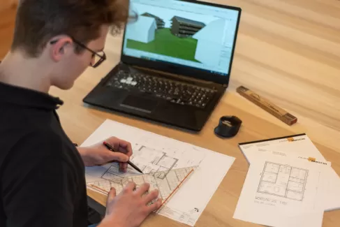 Ein junger Mann arbeitet konzentriert an einem Architektenarbeitsplatz. Vor ihm liegt ein detaillierter Grundrissplan, den er mit Lineal und Bleistift bearbeitet. Im Hintergrund läuft auf dem Laptop ein 3D-Modell eines Gebäudes. Auf dem Tisch liegen weitere Pläne, ein Bleistift, ein Gliedermaßstab und ein Mauspad mit dem Logo der Zimmerei Hirschi AG. Das Bild wirbt für eine Lehrstelle als Zeichner:in Fachrichtung Architektur EFZ ab August 2026.