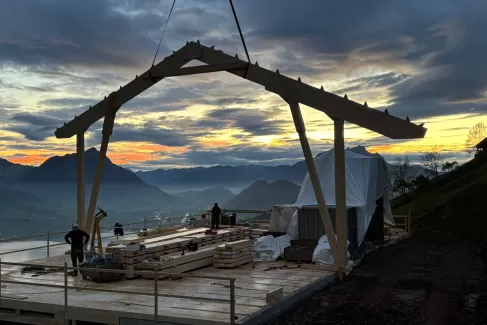 Holzbauarbeiten auf einer Bergbaustelle bei Sonnenaufgang – Zimmerleute montieren eine Dachkonstruktion mit Blick auf die umliegenden Berge und das Tal unter einem farbenprächtigen Morgenhimmel.