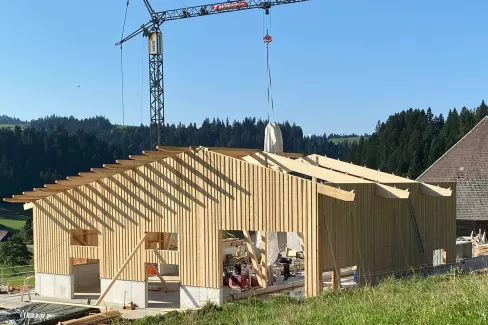 Ein modernes Holzbauprojekt von Zimmerei Hirschi AG: Im Vordergrund ist ein neu errichtetes Gebäude mit tragender Holzkonstruktion zu sehen. Ein Baukran hebt Materialien auf das Dach, während die klare Struktur der Holzbalken und Fassaden die Handwerkskunst hervorhebt. Umgeben von grünen Wiesen und bewaldeten Hügeln steht der Bau für nachhaltige, präzise und regionale Zimmermannsarbeit.
