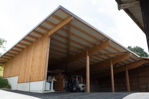 Aussenansicht einer grossen, offenen Lager- oder Maschinenhalle in Holzbauweise mit geneigtem Dach. Die Konstruktion aus sichtbaren Holzbalken ruht auf einem Betonsockel. Im Inneren der Halle sind ein Gabelstapler und gelagerte Materialien zu erkennen. Die Umgebung wirkt ländlich und funktional gestaltet.
