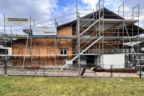 Holzhaus im Bau mit eingerüsteter Fassade und Baustellenbanner der Zimmerei Hirschi unter blauem Himmel.