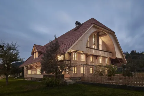 Traditionelles Holzhaus im Emmental in der Abenddämmerung, mit geschwungenem Giebeldach und aufwendig gestalteter Holzfassade. Warmes Licht fällt aus den Fenstern und betont die handwerklichen Details der historischen Holzkonstruktion. Das Gebäude steht in einer ruhigen, ländlichen Umgebung mit Garten, Bäumen und Holzzaun.