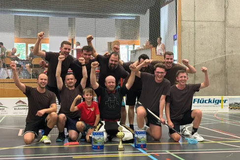 Eine Unihockey-Mannschaft feiert gemeinsam mit einem Kind und einem Torhüter auf dem Hallenboden einen Turniersieg. Vor der Gruppe stehen ein Pokal und zwei Getränkepreise.