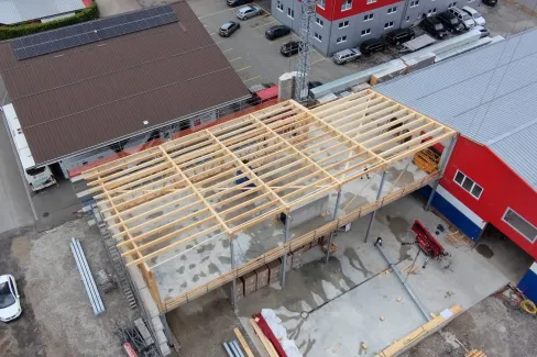 Luftaufnahme eines Gewerbebaus während der Bauphase. Zu sehen ist die im Aufbau befindliche Holzdachkonstruktion mit Tragwerk und Sparren auf einem Industriegebäude, umgeben von Werkhallen und Baustelleneinrichtungen.