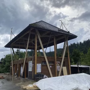 Modernes Holzgebäude mit markantem, erhöhtem Satteldach und offener Konstruktion, aufgenommen an einem bewölkten Tag inmitten einer grünen Hügellandschaft.
