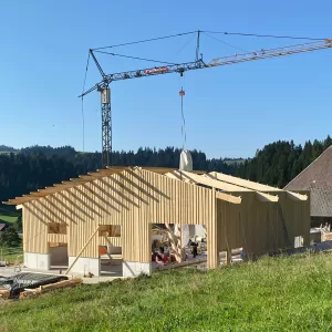 Ein modernes Holzbauprojekt von Zimmerei Hirschi AG: Im Vordergrund ist ein neu errichtetes Gebäude mit tragender Holzkonstruktion zu sehen. Ein Baukran hebt Materialien auf das Dach, während die klare Struktur der Holzbalken und Fassaden die Handwerkskunst hervorhebt. Umgeben von grünen Wiesen und bewaldeten Hügeln steht der Bau für nachhaltige, präzise und regionale Zimmermannsarbeit.