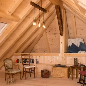 Gemütlicher Dachraum mit sichtbarer Holzbalkendecke, Holzboden und Einrichtung aus Stühlen, Bücherregal und warmem Licht.
