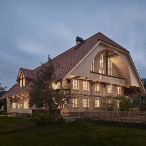 Traditionelles Holzhaus im Emmental in der Abenddämmerung, mit geschwungenem Giebeldach und aufwendig gestalteter Holzfassade. Warmes Licht fällt aus den Fenstern und betont die handwerklichen Details der historischen Holzkonstruktion. Das Gebäude steht in einer ruhigen, ländlichen Umgebung mit Garten, Bäumen und Holzzaun.