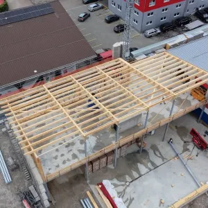 Luftaufnahme eines Gewerbebaus während der Bauphase. Zu sehen ist die im Aufbau befindliche Holzdachkonstruktion mit Tragwerk und Sparren auf einem Industriegebäude, umgeben von Werkhallen und Baustelleneinrichtungen.
