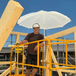 Innenansicht einer im Bau befindlichen Gewerbehalle mit weit gespannter Holzdachkonstruktion aus Brettschichtholz. Sichtbar sind die tragenden Binder, Sparren und die Dachschalung, während eine Hebebühne auf der Holzdecke den Baufortschritt der Umnutzung dokumentiert.