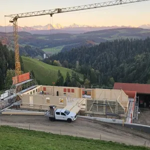 Holzbau im Rohbau mit Baukran an einem grünen Hang, mit bewaldeten Hügeln und schneebedeckten Bergen im Hintergrund.