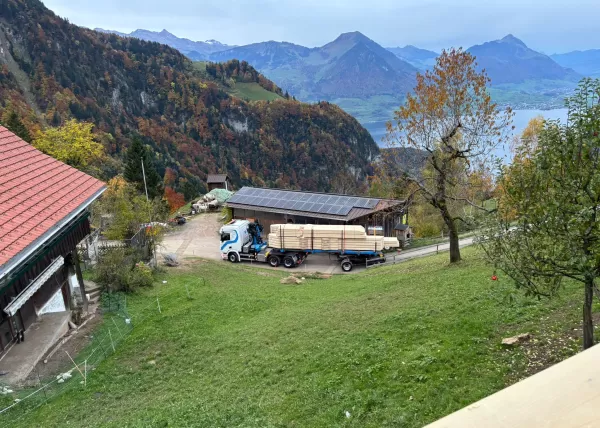 Ein Lastwagen mit Holzlieferung steht auf einem Hof inmitten einer herbstlichen Berglandschaft – im Hintergrund sind Wälder, Berge und ein See zu sehen.
