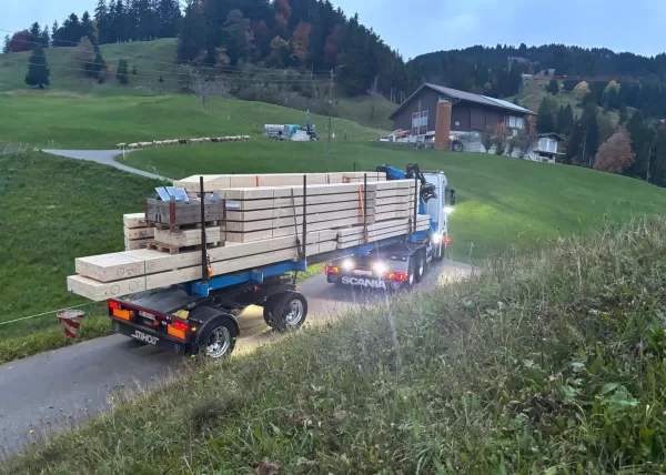 Ein Lastwagen der Zimmerei liefert große Holzbalken auf einer kurvigen Bergstraße, umgeben von grünen Wiesen und Höfen in hügeliger Landschaft.