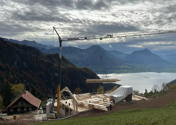 Auf einer Bergbaustelle hebt ein Kran Holzbalken über eine Holzbaukonstruktion mit Blick auf einen See und umliegende Berge unter bewölktem Himmel.