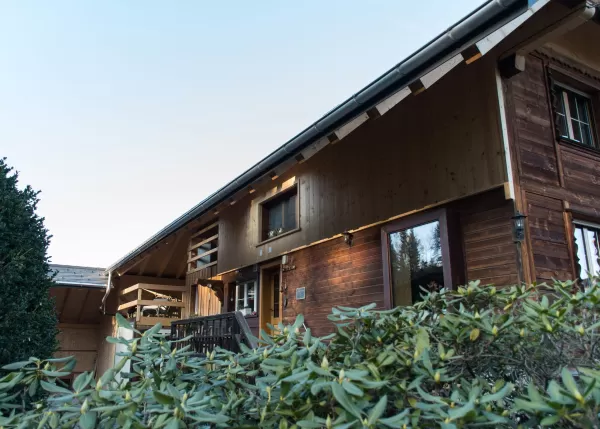 Holzhaus mit Balkon und Veranda, umgeben von grünen Sträuchern, bei klarem Himmel von außen aufgenommen.