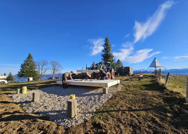 Montagearbeiten an einer Holzplattform vor einem traditionellen Gebäude auf einer Anhöhe. Ein Traktor mit Hebevorrichtung hebt einen langen Holzbalken an, während zwei Mitarbeitende der Zimmerei Hirschi AG den Balken ausrichten.