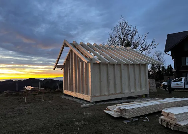Montagearbeiten an einer Holzplattform vor einem traditionellen Gebäude auf einer Anhöhe. Ein Traktor mit Hebevorrichtung hebt einen langen Holzbalken an, während zwei Mitarbeitende der Zimmerei Hirschi AG den Balken ausrichten.