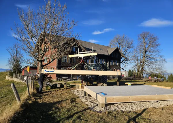 Montagearbeiten an einer Holzplattform vor einem traditionellen Gebäude auf einer Anhöhe. Ein Traktor mit Hebevorrichtung hebt einen langen Holzbalken an, während zwei Mitarbeitende der Zimmerei Hirschi AG den Balken ausrichten.