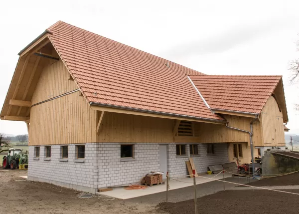 Neu erbautes landwirtschaftliches Gebäude mit Kombination aus Holzfassade und gemauertem Sockelgeschoss. Das rote Ziegeldach und die sichtbare Holzstruktur zeigen präzise Zimmermannsarbeit und traditionelle Bauweise in modernem Stil.