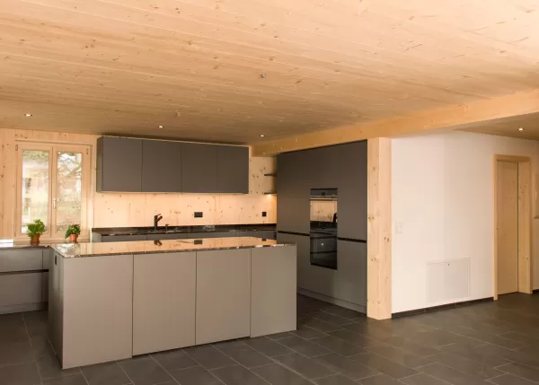 Moderne Küche mit grauen Fronten und Arbeitsplatte aus Stein, umgeben von hellen Holzwänden und -decke. Das harmonische Zusammenspiel von Holz und modernen Materialien schafft eine warme und zeitgemäße Wohnatmosphäre.