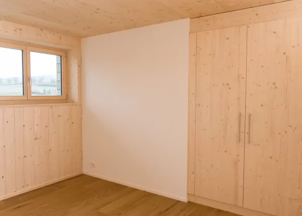 Helles Zimmer mit Holzdecke, Wandverkleidung und Einbauschrank aus Fichtenholz. Ein grosses Fenster sorgt für natürliches Licht und betont die warme, natürliche Raumgestaltung.