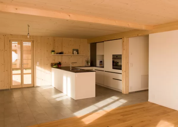 Offene, helle Wohnküche mit modernem weißen Küchenblock und Holzverkleidung. Die Kombination aus Holzdecke, Fliesen- und Parkettboden schafft ein natürliches und einladendes Raumgefühl.