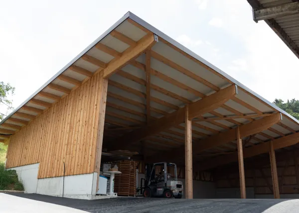 Aussenansicht einer grossen, offenen Lager- oder Maschinenhalle in Holzbauweise mit geneigtem Dach. Die Konstruktion aus sichtbaren Holzbalken ruht auf einem Betonsockel. Im Inneren der Halle sind ein Gabelstapler und gelagerte Materialien zu erkennen. Die Umgebung wirkt ländlich und funktional gestaltet.