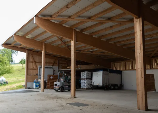 Aussenansicht einer grossen, offenen Lager- oder Maschinenhalle in Holzbauweise mit geneigtem Dach. Die Konstruktion aus sichtbaren Holzbalken ruht auf einem Betonsockel. Im Inneren der Halle sind ein Gabelstapler und gelagerte Materialien zu erkennen. Die Umgebung wirkt ländlich und funktional gestaltet.