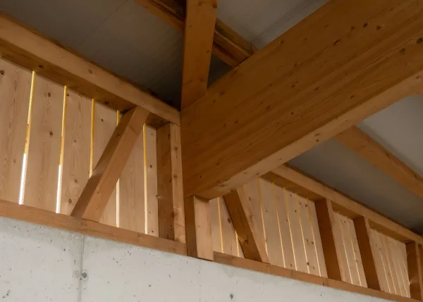 Aussenansicht einer grossen, offenen Lager- oder Maschinenhalle in Holzbauweise mit geneigtem Dach. Die Konstruktion aus sichtbaren Holzbalken ruht auf einem Betonsockel. Im Inneren der Halle sind ein Gabelstapler und gelagerte Materialien zu erkennen. Die Umgebung wirkt ländlich und funktional gestaltet.