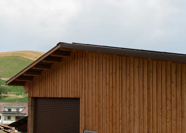 Aussenansicht einer grossen, offenen Lager- oder Maschinenhalle in Holzbauweise mit geneigtem Dach. Die Konstruktion aus sichtbaren Holzbalken ruht auf einem Betonsockel. Im Inneren der Halle sind ein Gabelstapler und gelagerte Materialien zu erkennen. Die Umgebung wirkt ländlich und funktional gestaltet.