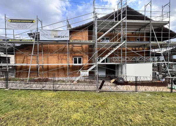 Holzhaus im Bau mit eingerüsteter Fassade und Baustellenbanner der Zimmerei Hirschi unter blauem Himmel.