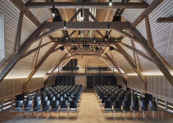 Traditionelles Holzhaus im Emmental in der Abenddämmerung, mit geschwungenem Giebeldach und aufwendig gestalteter Holzfassade. Warmes Licht fällt aus den Fenstern und betont die handwerklichen Details der historischen Holzkonstruktion. Das Gebäude steht in einer ruhigen, ländlichen Umgebung mit Garten, Bäumen und Holzzaun.