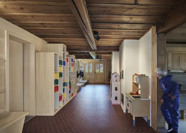 Traditionelles Holzhaus im Emmental in der Abenddämmerung, mit geschwungenem Giebeldach und aufwendig gestalteter Holzfassade. Warmes Licht fällt aus den Fenstern und betont die handwerklichen Details der historischen Holzkonstruktion. Das Gebäude steht in einer ruhigen, ländlichen Umgebung mit Garten, Bäumen und Holzzaun.