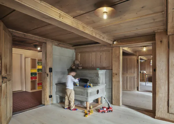 Traditionelles Holzhaus im Emmental in der Abenddämmerung, mit geschwungenem Giebeldach und aufwendig gestalteter Holzfassade. Warmes Licht fällt aus den Fenstern und betont die handwerklichen Details der historischen Holzkonstruktion. Das Gebäude steht in einer ruhigen, ländlichen Umgebung mit Garten, Bäumen und Holzzaun.