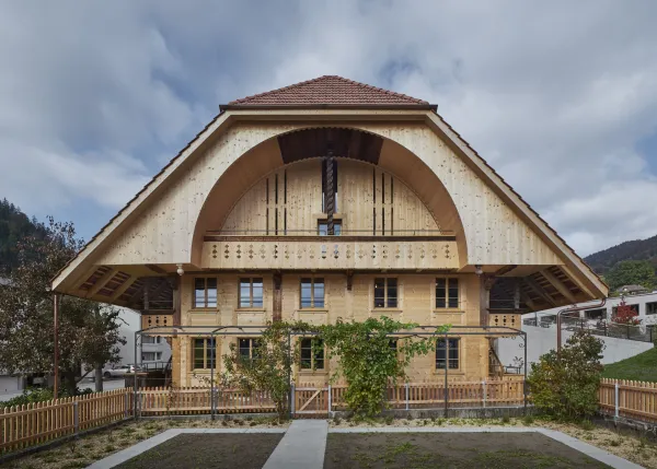 Traditionelles Holzhaus im Emmental in der Abenddämmerung, mit geschwungenem Giebeldach und aufwendig gestalteter Holzfassade. Warmes Licht fällt aus den Fenstern und betont die handwerklichen Details der historischen Holzkonstruktion. Das Gebäude steht in einer ruhigen, ländlichen Umgebung mit Garten, Bäumen und Holzzaun.