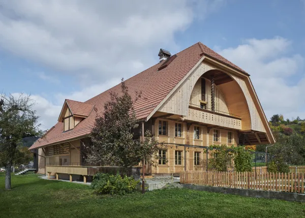Traditionelles Holzhaus im Emmental in der Abenddämmerung, mit geschwungenem Giebeldach und aufwendig gestalteter Holzfassade. Warmes Licht fällt aus den Fenstern und betont die handwerklichen Details der historischen Holzkonstruktion. Das Gebäude steht in einer ruhigen, ländlichen Umgebung mit Garten, Bäumen und Holzzaun.