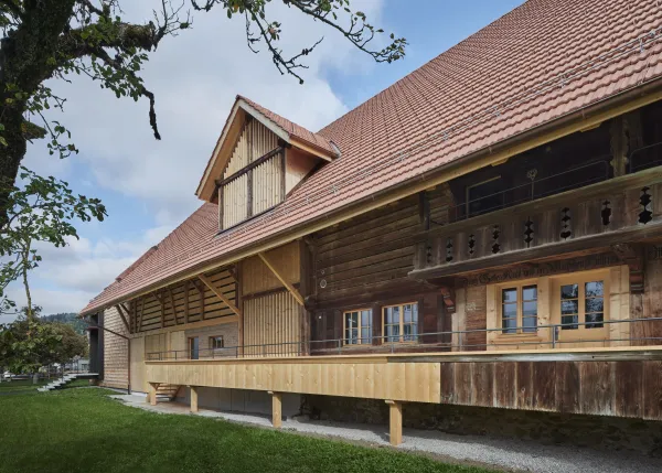 Traditionelles Holzhaus im Emmental in der Abenddämmerung, mit geschwungenem Giebeldach und aufwendig gestalteter Holzfassade. Warmes Licht fällt aus den Fenstern und betont die handwerklichen Details der historischen Holzkonstruktion. Das Gebäude steht in einer ruhigen, ländlichen Umgebung mit Garten, Bäumen und Holzzaun.
