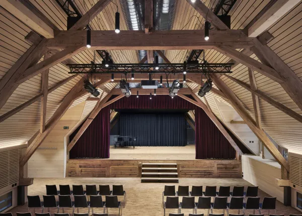 Traditionelles Holzhaus im Emmental in der Abenddämmerung, mit geschwungenem Giebeldach und aufwendig gestalteter Holzfassade. Warmes Licht fällt aus den Fenstern und betont die handwerklichen Details der historischen Holzkonstruktion. Das Gebäude steht in einer ruhigen, ländlichen Umgebung mit Garten, Bäumen und Holzzaun.