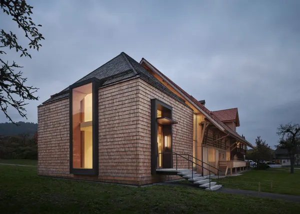 Traditionelles Holzhaus im Emmental in der Abenddämmerung, mit geschwungenem Giebeldach und aufwendig gestalteter Holzfassade. Warmes Licht fällt aus den Fenstern und betont die handwerklichen Details der historischen Holzkonstruktion. Das Gebäude steht in einer ruhigen, ländlichen Umgebung mit Garten, Bäumen und Holzzaun.