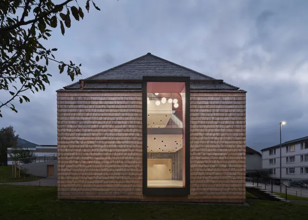 Traditionelles Holzhaus im Emmental in der Abenddämmerung, mit geschwungenem Giebeldach und aufwendig gestalteter Holzfassade. Warmes Licht fällt aus den Fenstern und betont die handwerklichen Details der historischen Holzkonstruktion. Das Gebäude steht in einer ruhigen, ländlichen Umgebung mit Garten, Bäumen und Holzzaun.