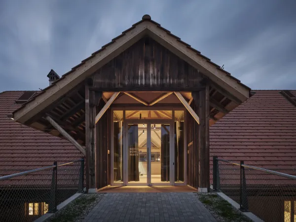 Traditionelles Holzhaus im Emmental in der Abenddämmerung, mit geschwungenem Giebeldach und aufwendig gestalteter Holzfassade. Warmes Licht fällt aus den Fenstern und betont die handwerklichen Details der historischen Holzkonstruktion. Das Gebäude steht in einer ruhigen, ländlichen Umgebung mit Garten, Bäumen und Holzzaun.