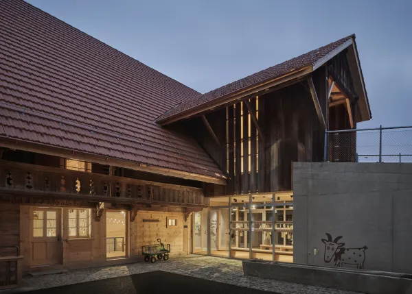 Traditionelles Holzhaus im Emmental in der Abenddämmerung, mit geschwungenem Giebeldach und aufwendig gestalteter Holzfassade. Warmes Licht fällt aus den Fenstern und betont die handwerklichen Details der historischen Holzkonstruktion. Das Gebäude steht in einer ruhigen, ländlichen Umgebung mit Garten, Bäumen und Holzzaun.
