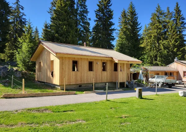 Holzhaus im Bau mit natürlicher Holzfassade inmitten einer Waldlandschaft. Das eingeschossige Gebäude steht auf einem gepflegten Grundstück mit Wiese und Zufahrtsweg. Die Fensteröffnungen sind bereits erstellt, während der Innenausbau noch im Gange ist. Im Hintergrund befinden sich weitere Holzbauten sowie hohe Nadelbäume, die das Gebäude umgeben. Die Aufnahme zeigt die harmonische Einbindung des Holzbaus in die natürliche Umgebung.