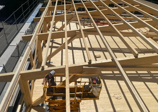 Innenansicht einer im Bau befindlichen Gewerbehalle mit weit gespannter Holzdachkonstruktion aus Brettschichtholz. Sichtbar sind die tragenden Binder, Sparren und die Dachschalung, während eine Hebebühne auf der Holzdecke den Baufortschritt der Umnutzung dokumentiert.